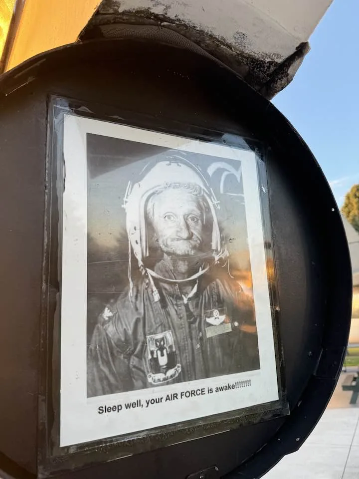 A black-and-white photo of a man in a military flight helmet and jacket is taped inside a public trash can. The photo has…