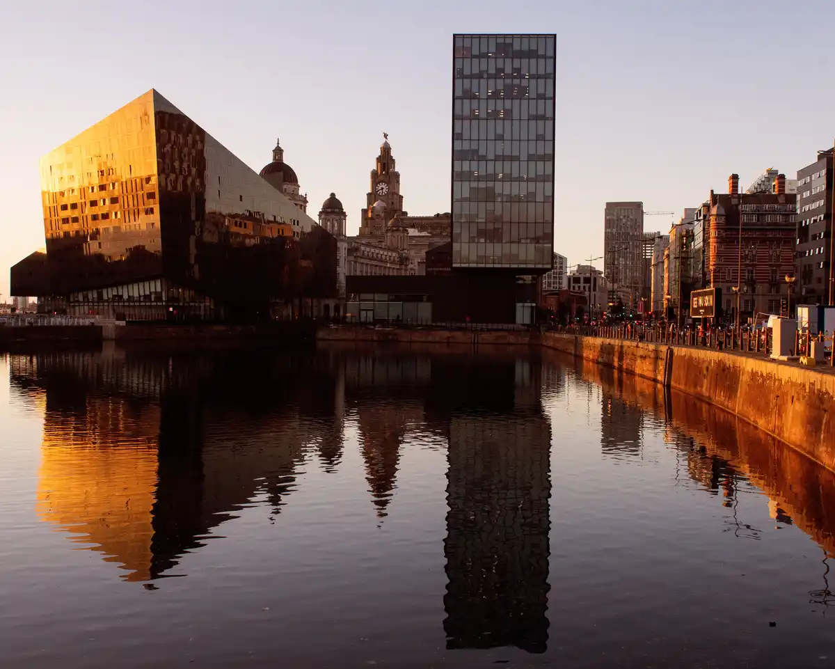A scenic photo of Liverpool's waterfront at sunset, featuring the modern Museum of Liverpool with its golden reflective…