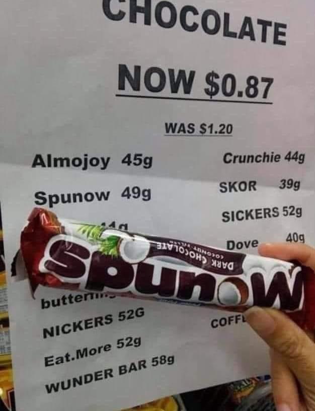 A hand holds a Spunow chocolate bar in front of a price list showing various chocolate bars and their weights. The list…