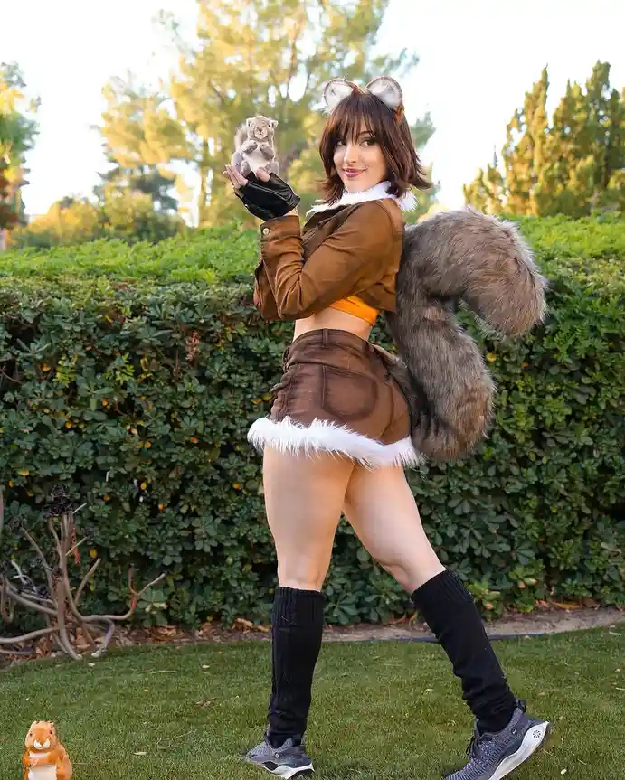 A woman in a detailed fox costume, complete with ears, a large bushy tail, and furry trim, poses outdoors holding a small…