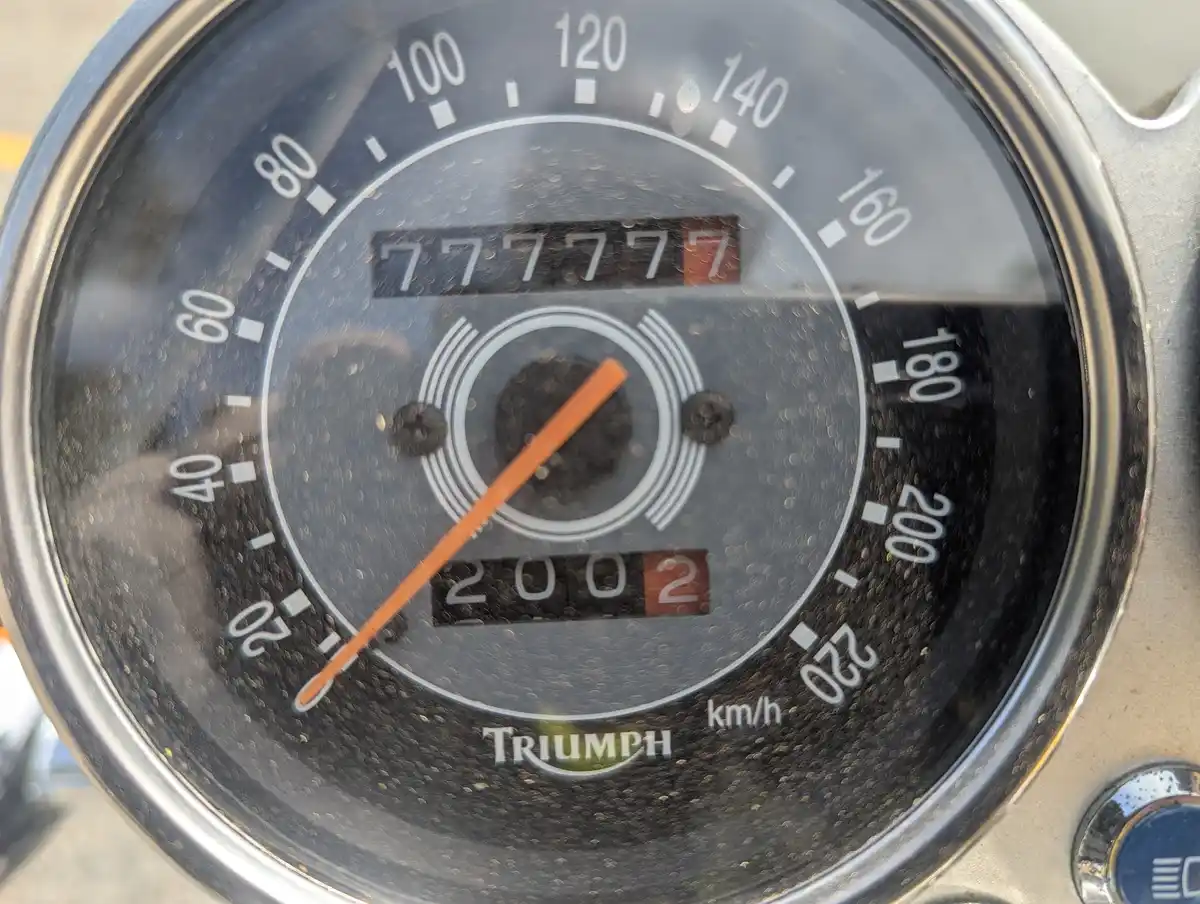 A close-up of a Triumph motorcycle speedometer showing the odometer reading '777777' and the speed at '0' km/h. The orange…