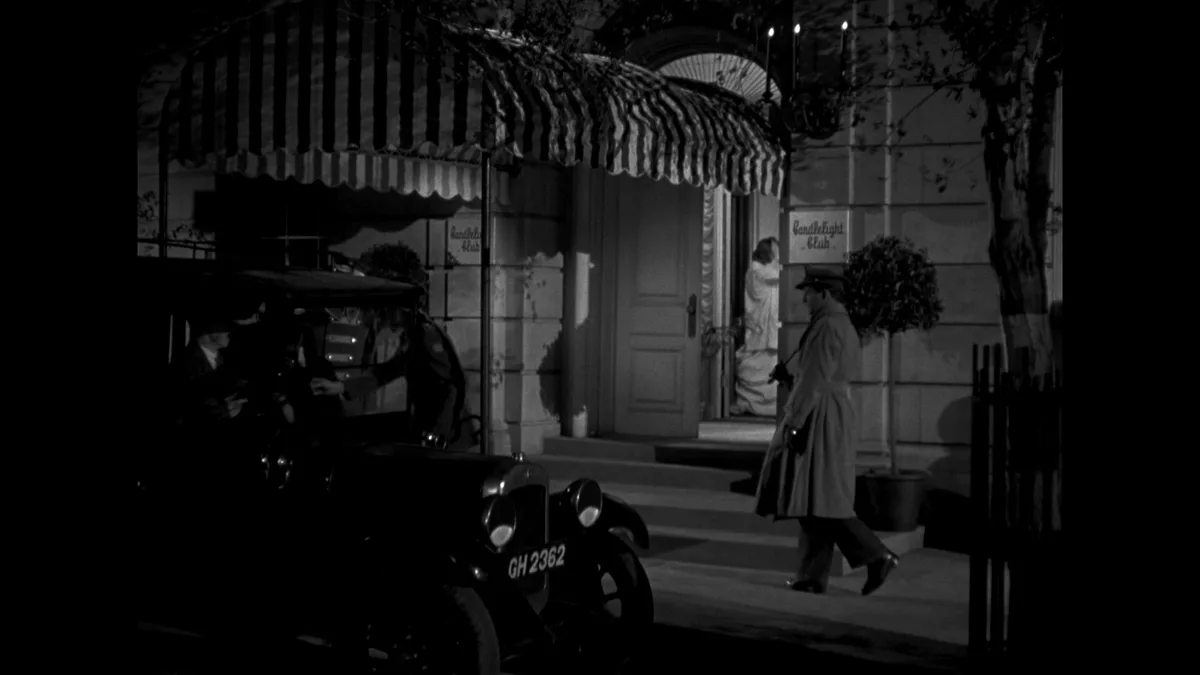 A black-and-white film still shows a man in a trench coat walking past a building entrance with a striped awning. A vintage…