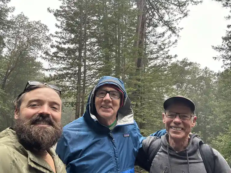 Three smiling men in rain gear pose for a selfie in a misty forest with tall trees behind them; the central man wears a blue…