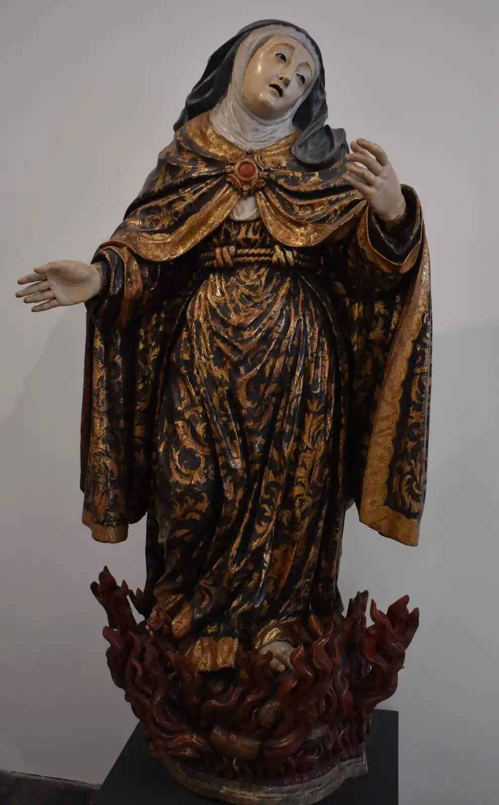 A wooden statue of a robed woman with a black veil, standing on a base carved to look like flames, with overlaid text…