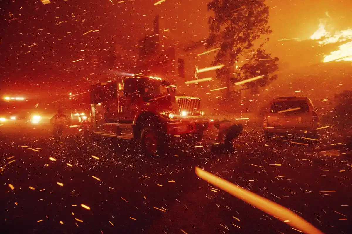 A dramatic nighttime photo shows a red fire truck parked on a street during a wildfire, with embers and sparks flying…