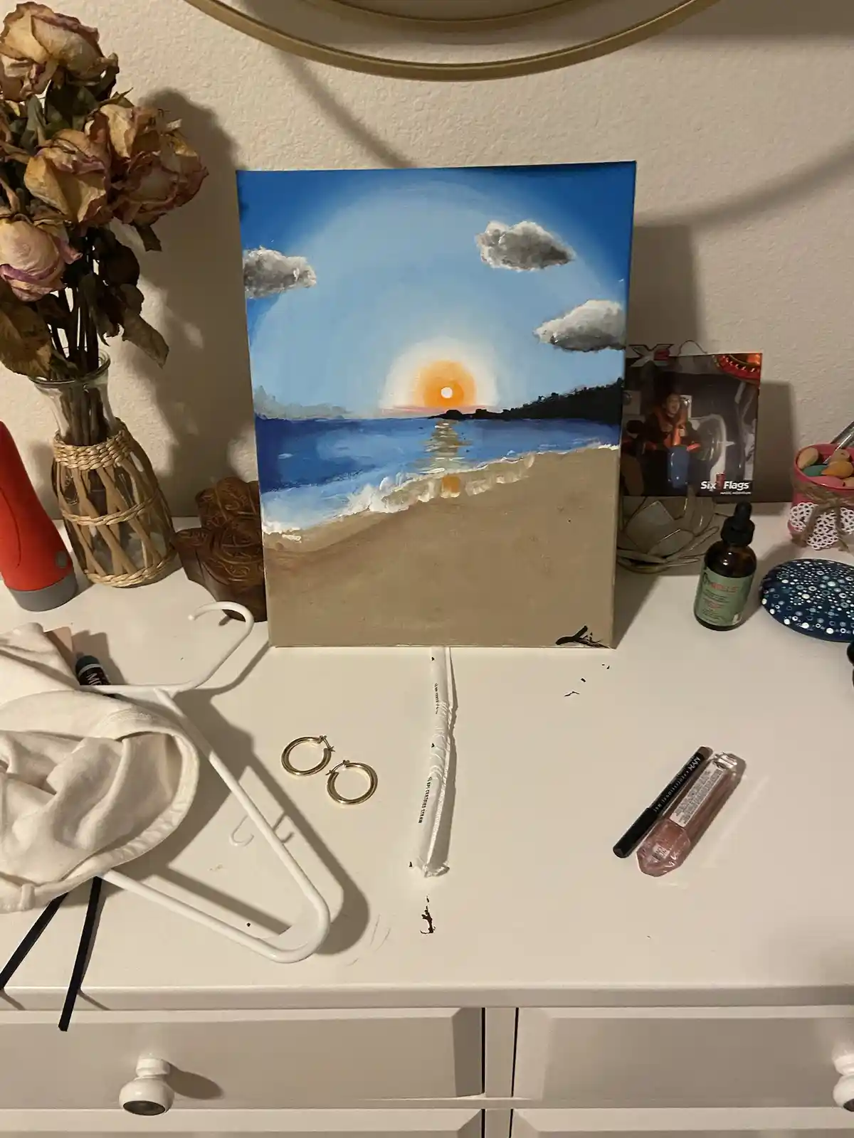 A handmade painting of a sunset over a beach sits upright on a white dresser, surrounded by everyday items like dried…