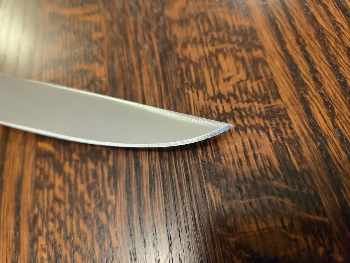 A close-up photo shows the sharp, metallic tip of a knife blade resting on a dark, textured wooden surface. The overlaid…