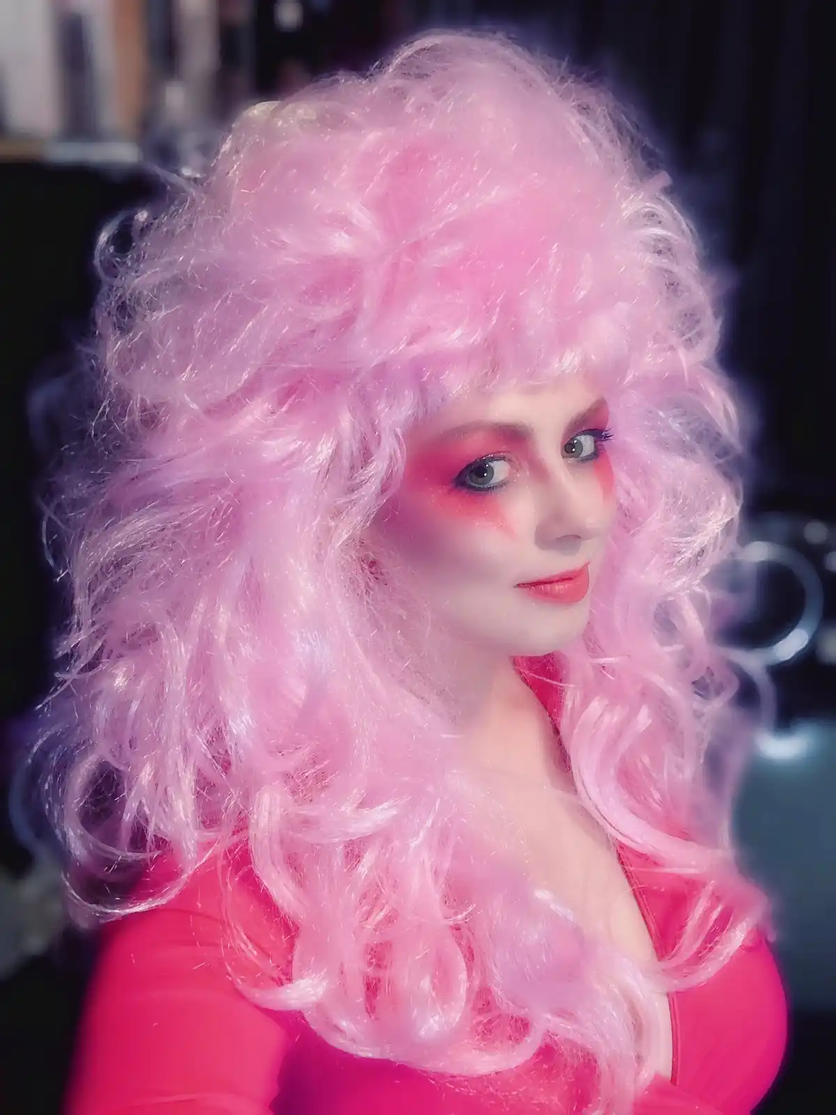 A person with a voluminous, curly pink wig and matching pink makeup and outfit looks directly at the camera with a neutral…