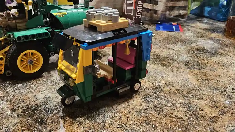 A small, colorful LEGO tuk-tuk with pink seats and a black roof sits on a speckled countertop, next to a larger green LEGO…