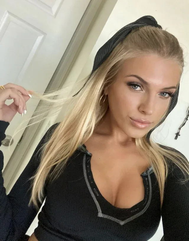 A blonde woman with blue eyes, wearing a black headband and black top, looks at the camera while holding a strand of her…