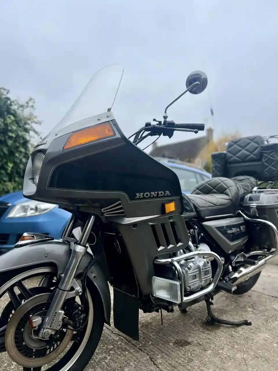 A grey Honda Goldwing motorcycle is parked outdoors, showing its front fairing, handlebars, and side-mounted saddlebags. The…