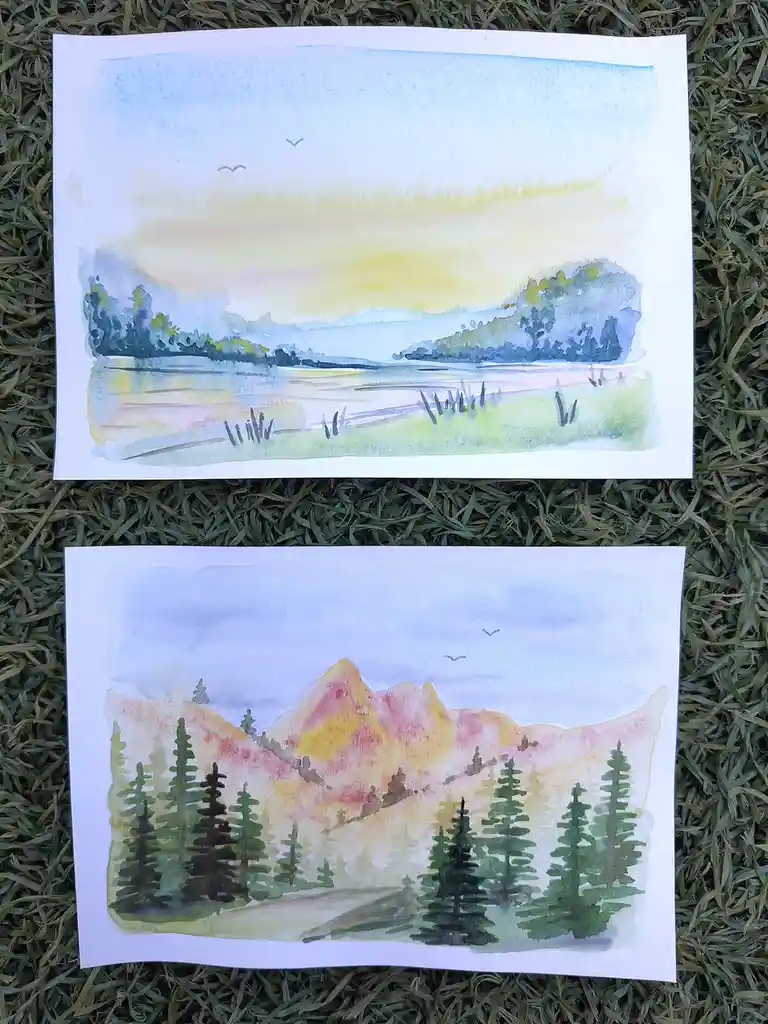 Two watercolor paintings lie on grass: the top one depicts a serene lake with trees and birds under a soft sky, while the…