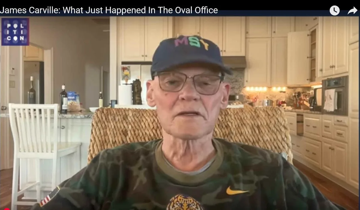 An older man with glasses and a blue cap with 'MSY' in colorful letters sits in a kitchen chair, wearing a camouflage shirt…