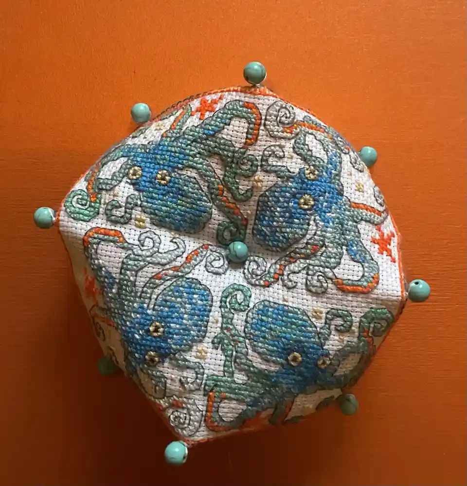 A hand-embroidered, hexagonal pin cushion with turquoise beads at each corner, featuring intricate blue and green octopus…