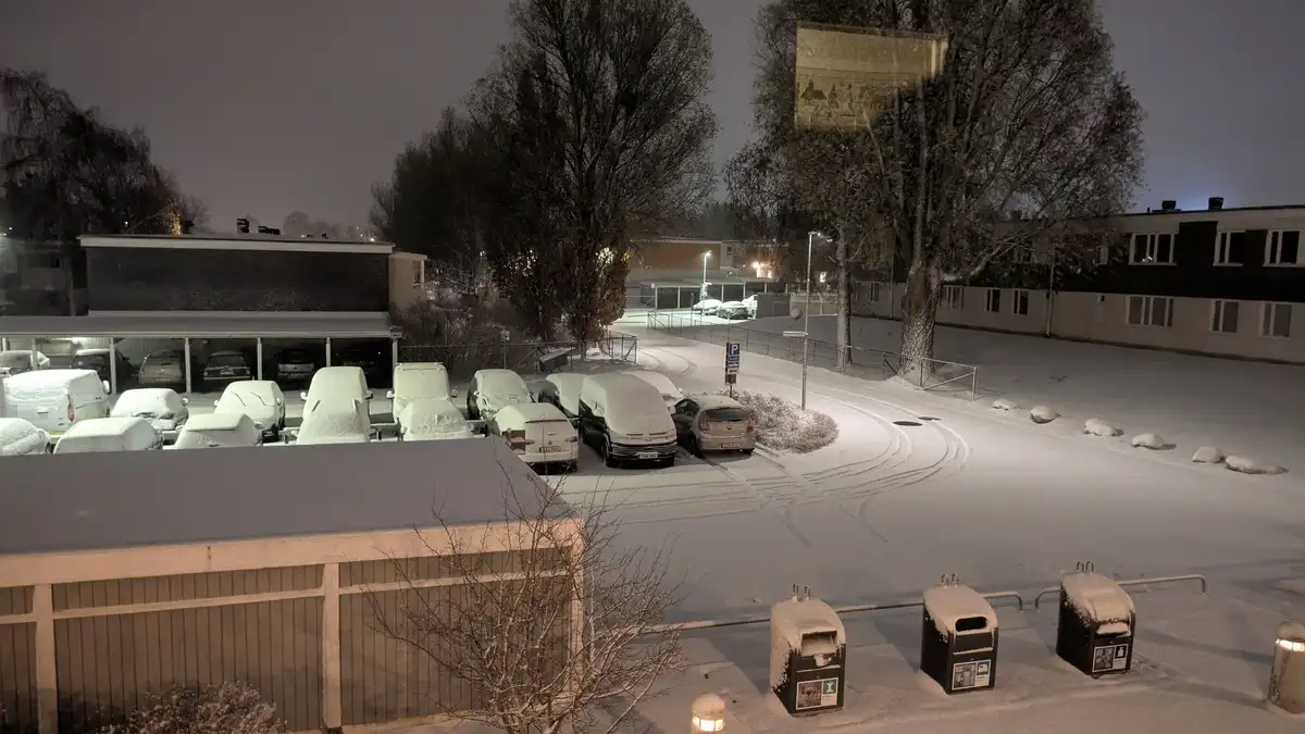 A nighttime photo of a snow-covered parking lot with cars buried under deep snow, showing tire tracks in the snow and…