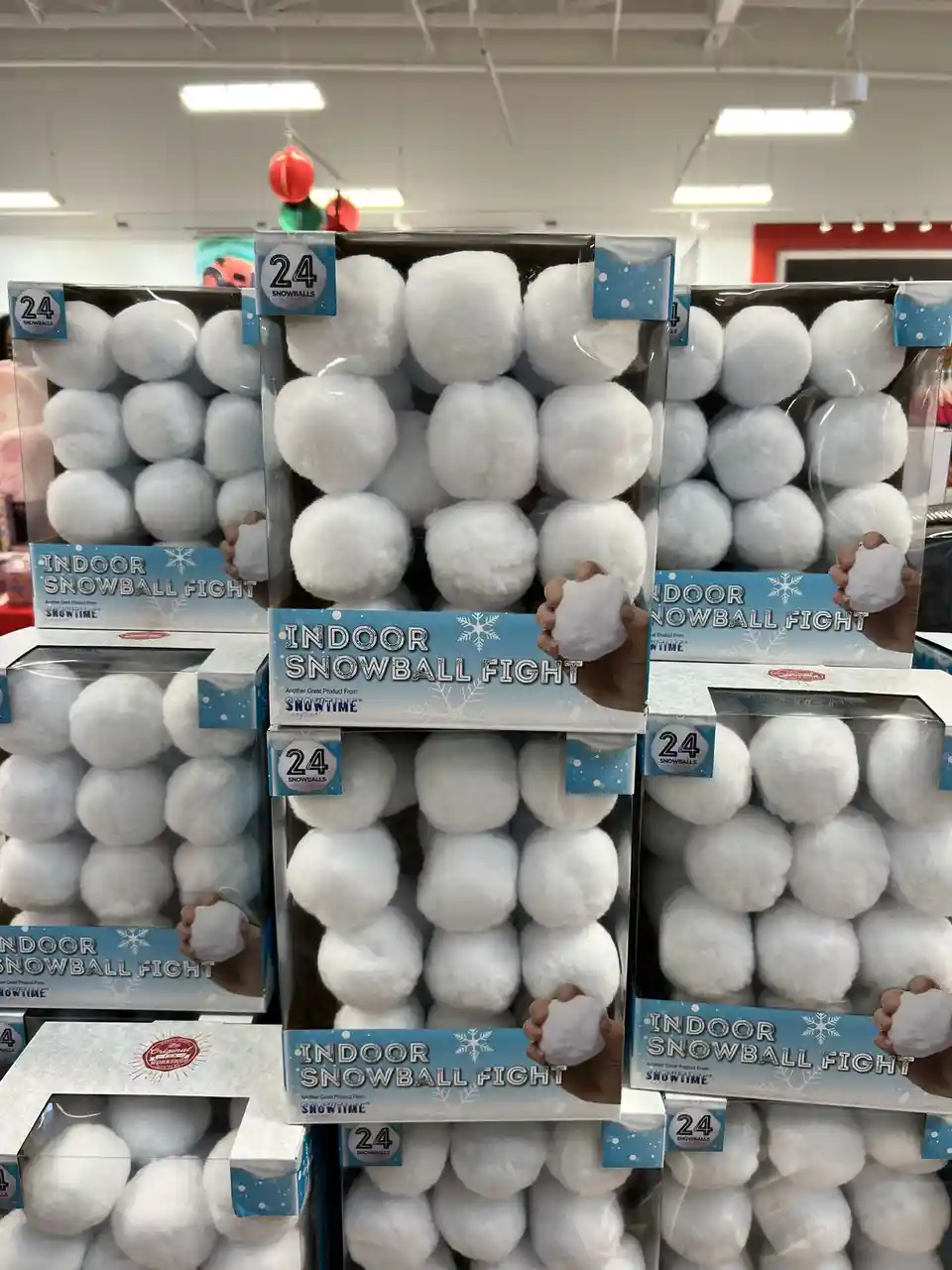 A store shelf displays multiple boxes of 'Indoor Snowball Fight' toys, each containing 24 fluffy white foam snowballs. The…