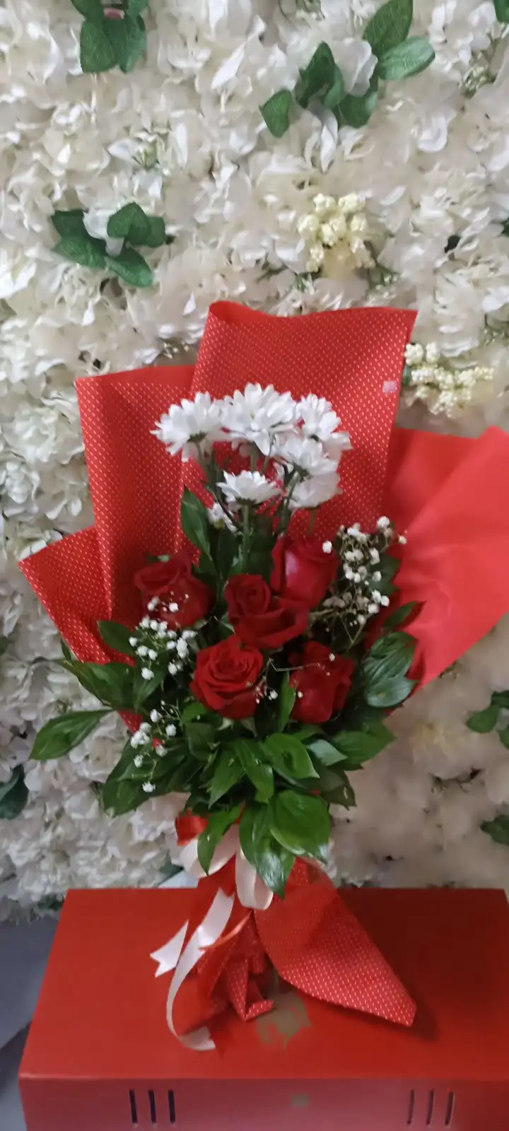 A bouquet of red roses and white daisies wrapped in red paper with a red and white ribbon sits on a red box, against a…