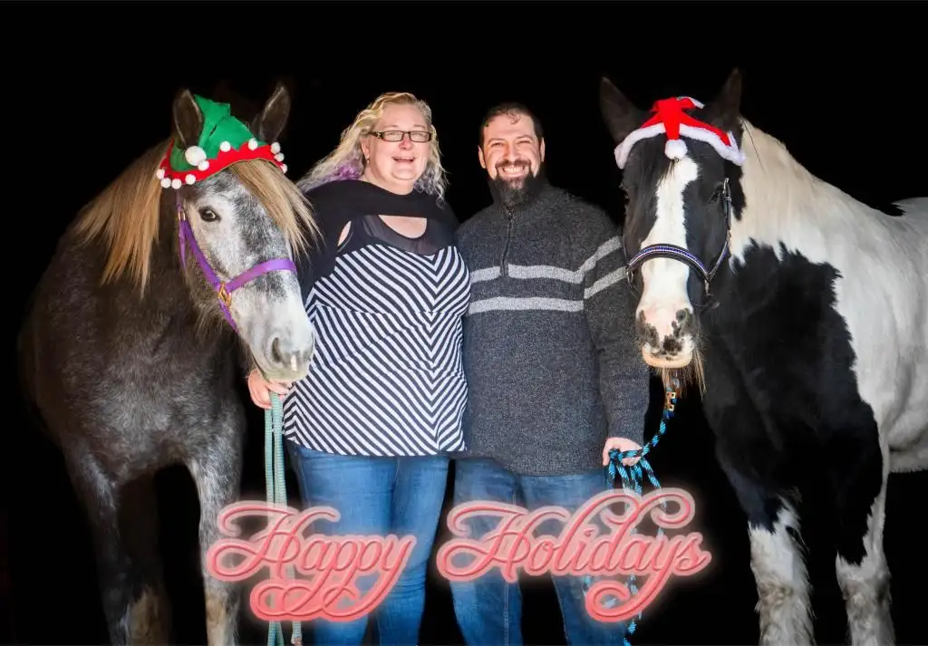 A man and woman stand smiling between two horses against a black background, each horse wearing a festive holiday hat — one…