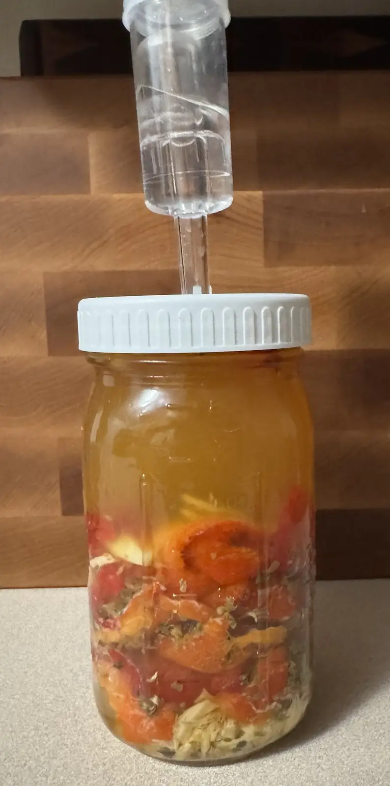 A glass mason jar filled with orange and red fruit pieces and herbs sits on a countertop, topped with a white lid and a…