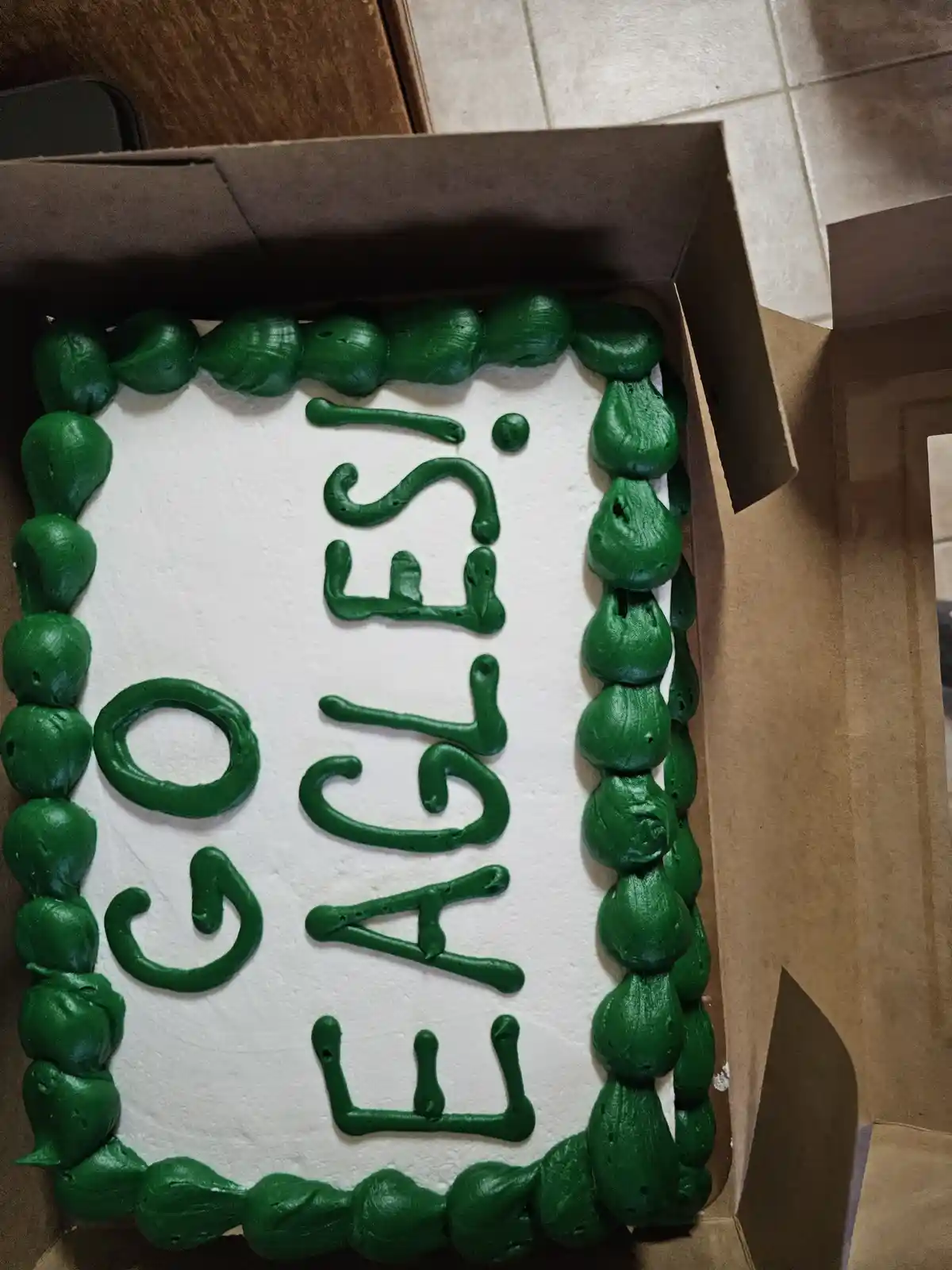 A rectangular cake in a cardboard box has white frosting with green piped icing spelling 'IS IT CAKE' vertically in the…