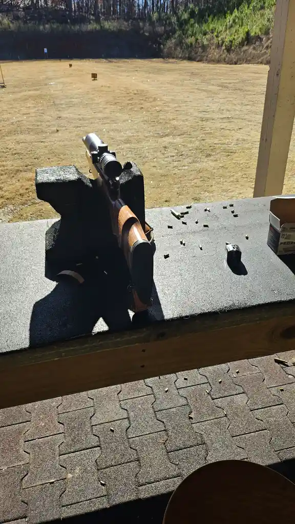 A rifle with a scope is mounted on a shooting bench at an outdoor range, aimed at a distant target marked '29'. Scattered…