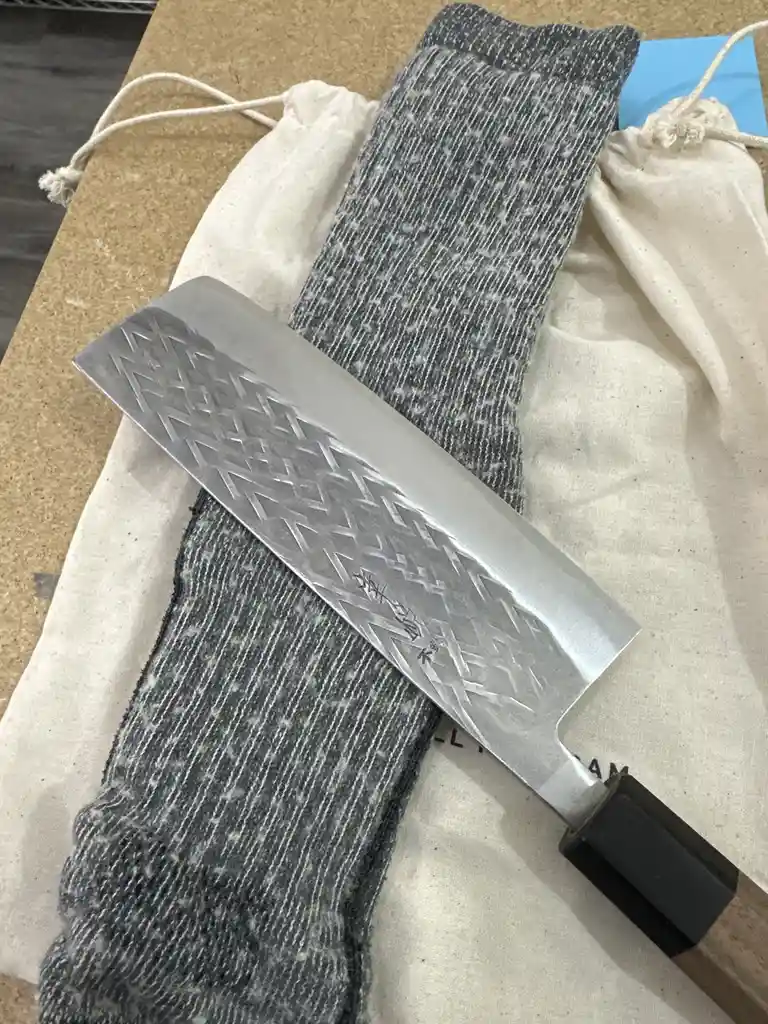 A large, ornate chef's knife with a patterned blade rests diagonally across a grey sock and a white drawstring bag on a…