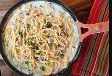 A close-up photo of a creamy spaghetti carbonara dish in a black skillet with a pink handle, resting on a colorful striped…