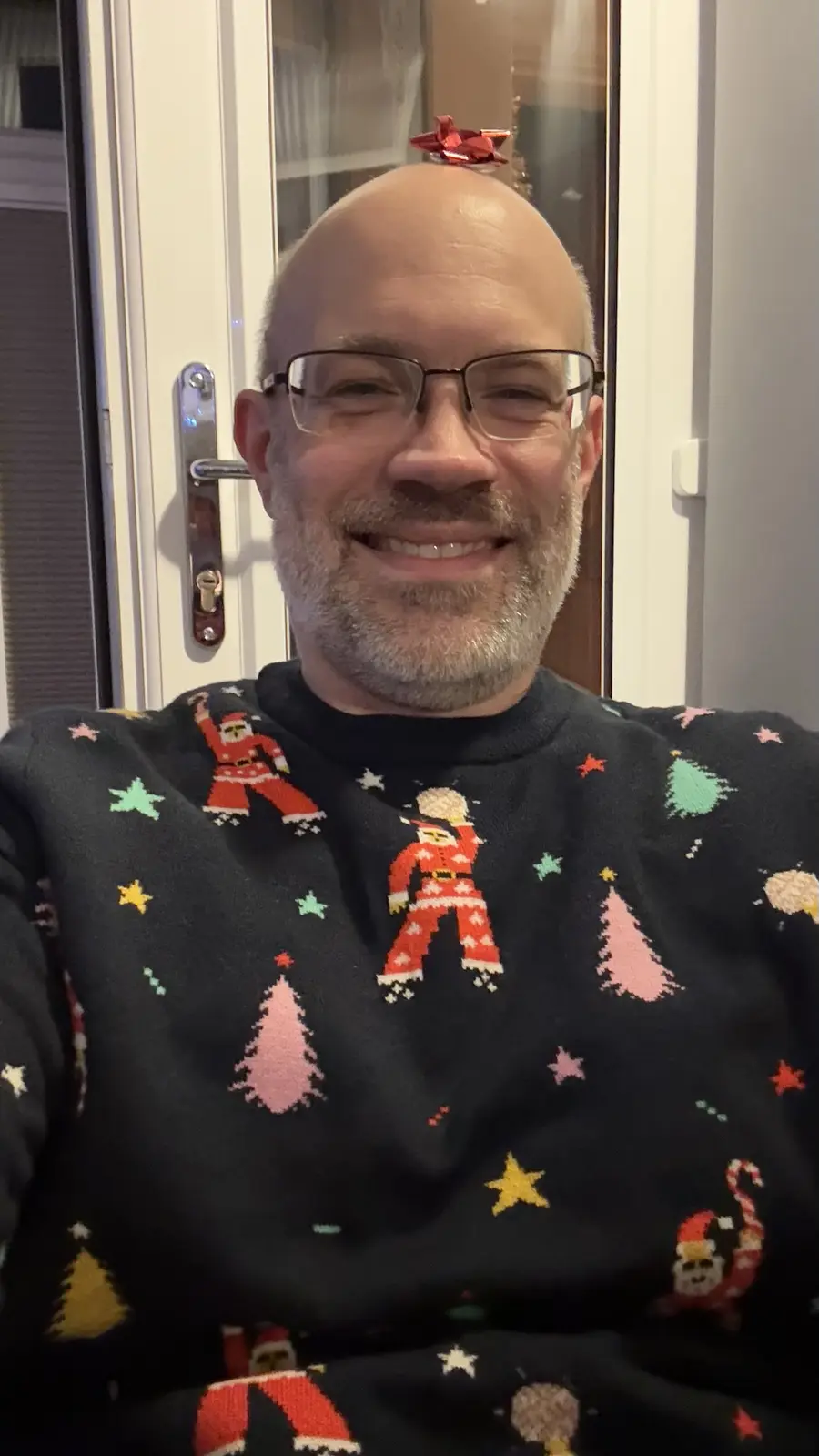 A bald man with glasses and a gray beard smiles at the camera while wearing a dark Christmas sweater covered in pixelated…