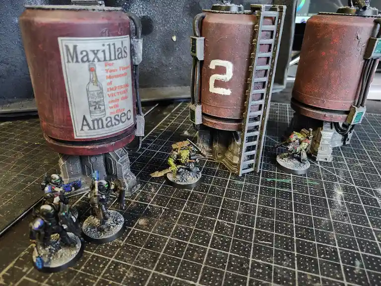 Three large, rusted, cylindrical tanks sit on a grid-patterned gaming mat, one labeled 'Maxilla's Amasec' with a slogan…