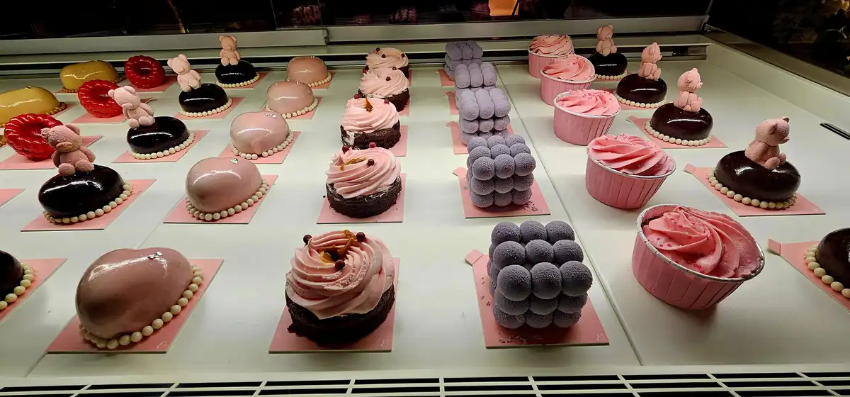 A display case filled with various pastries, including pink and chocolate domes, cupcakes with pink frosting, and purple…