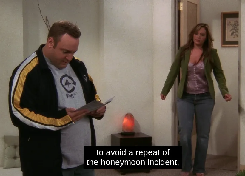 A man in a black and yellow jacket reads a letter while a woman in a green jacket walks into the room behind him. A glowing…