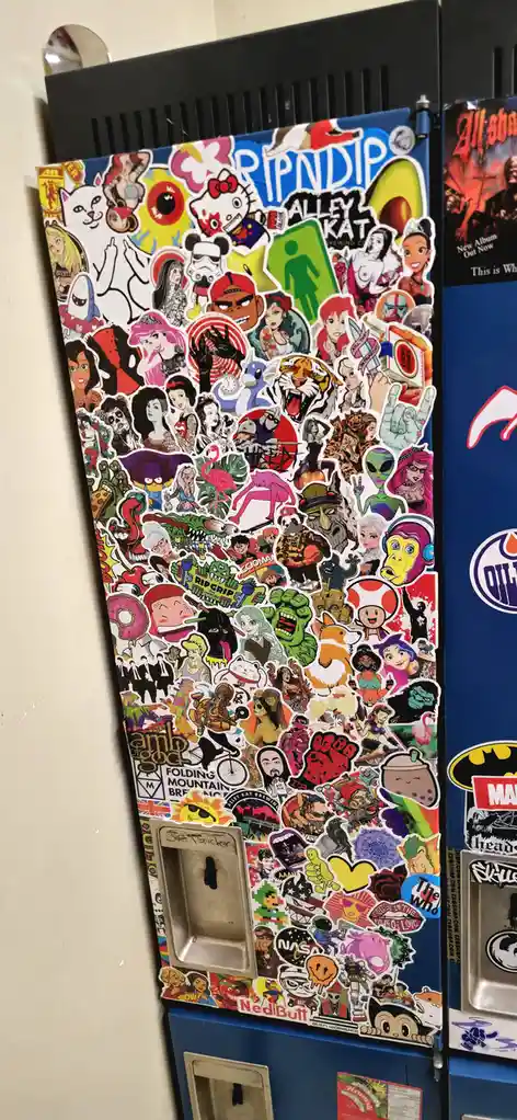 A blue locker door is completely covered in a dense, colorful collage of hundreds of overlapping stickers featuring cartoon…