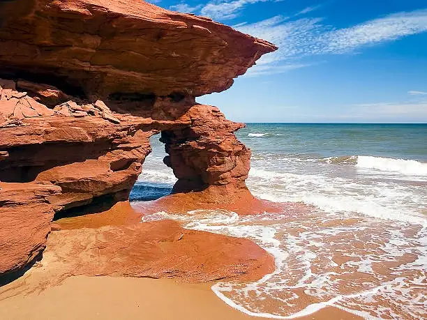 A scenic photo of a red sandstone sea arch formation on a beach with ocean waves crashing nearby under a blue sky. Overlaid…