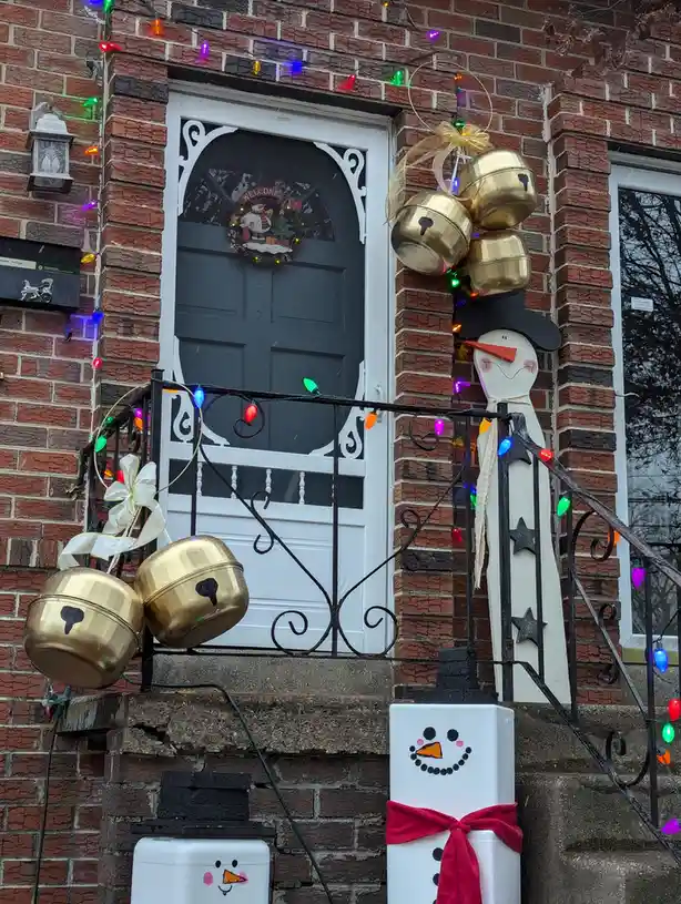 A festive brick house entrance decorated with Christmas lights, a wreath on the door, and two snowman figures made from…
