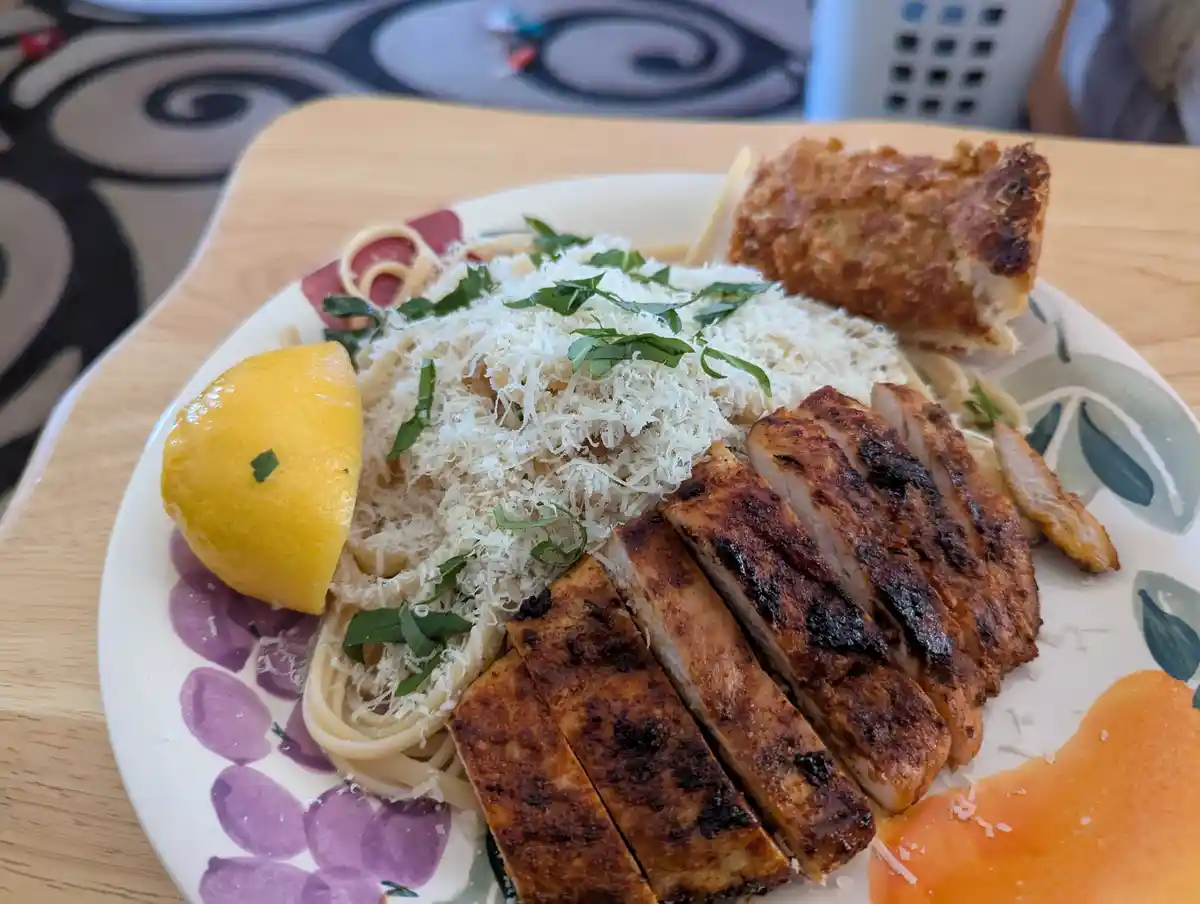 A plate of pasta topped with grated cheese and fresh basil, accompanied by sliced grilled chicken, a lemon wedge, and a…