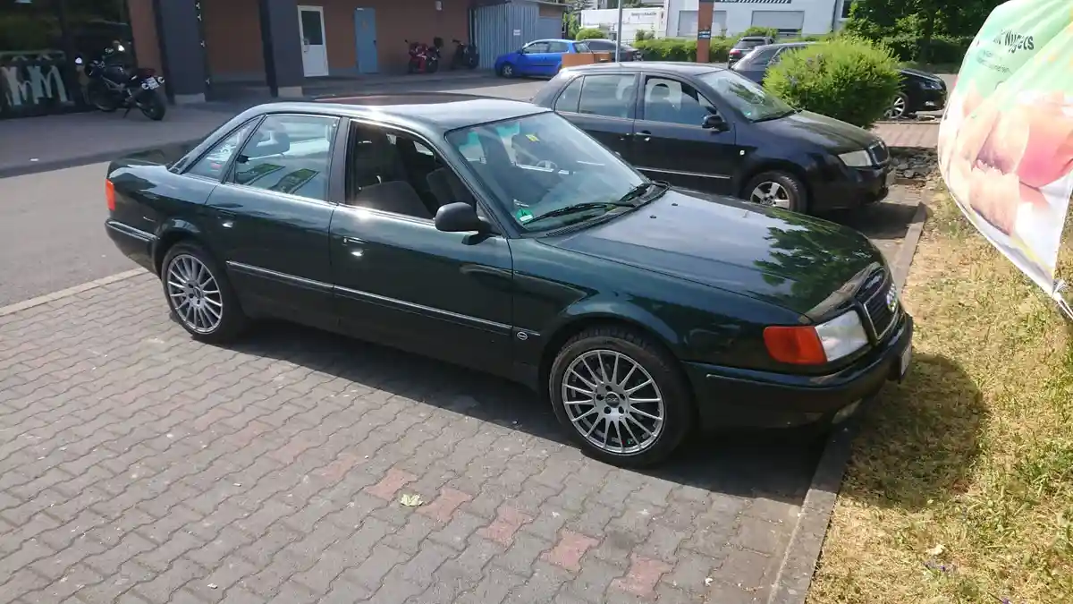 A dark green Audi A8 sedan is parked on a paved brick lot next to a grassy area with a promotional banner. The car has…