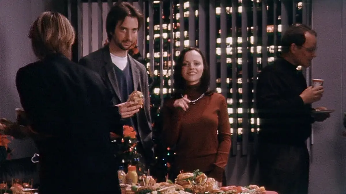 A still from a holiday party scene shows a woman in a brown turtleneck smiling and pointing at someone off-screen while…