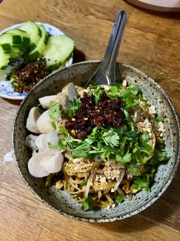 A close-up photo of a bowl of noodle dish topped with cilantro, crushed peanuts, and a large pile of dark red chili oil…