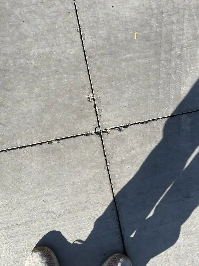 A top-down view of a person's feet in white sneakers standing on cracked concrete pavement, with their shadow cast across…
