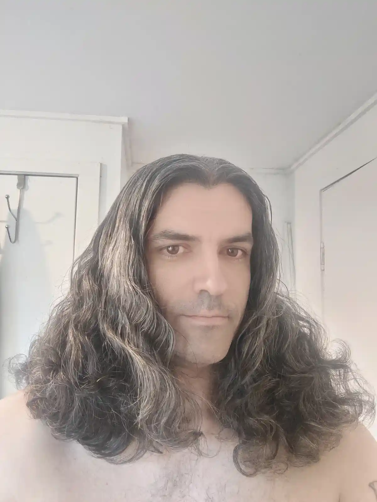 A shirtless man with long, wavy, dark hair with gray streaks stares directly at the camera with a neutral expression. The…