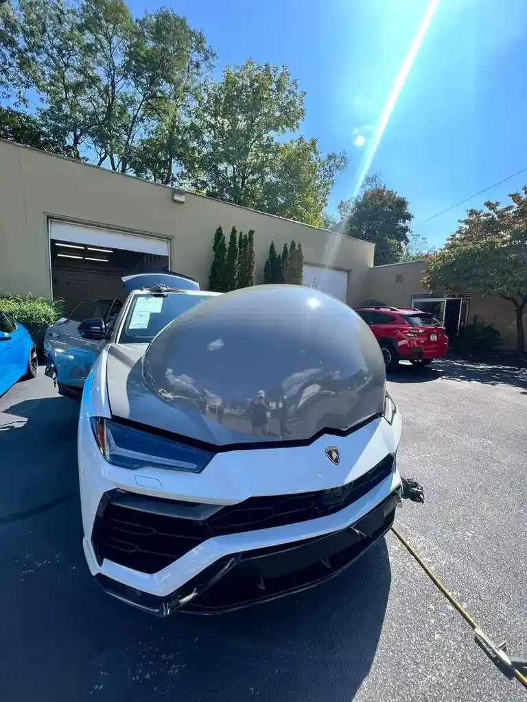 A white Lamborghini Urus is parked outdoors on a sunny day, its glossy hood reflecting the surroundings. The car's front…