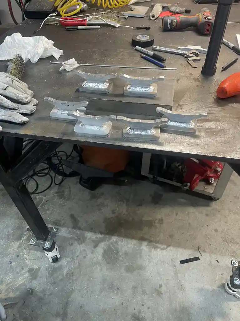 A cluttered workshop workbench displays six identical metal brackets, each with a welded seam, arranged in two rows…