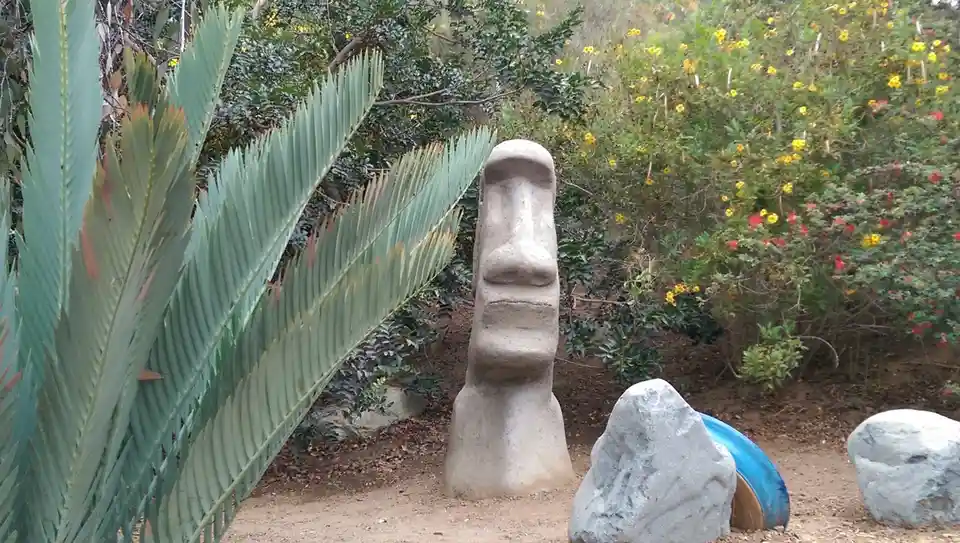 A gray Moai statue stands in a garden with green foliage and colorful flowers, with a blue plastic hat lying on the ground…