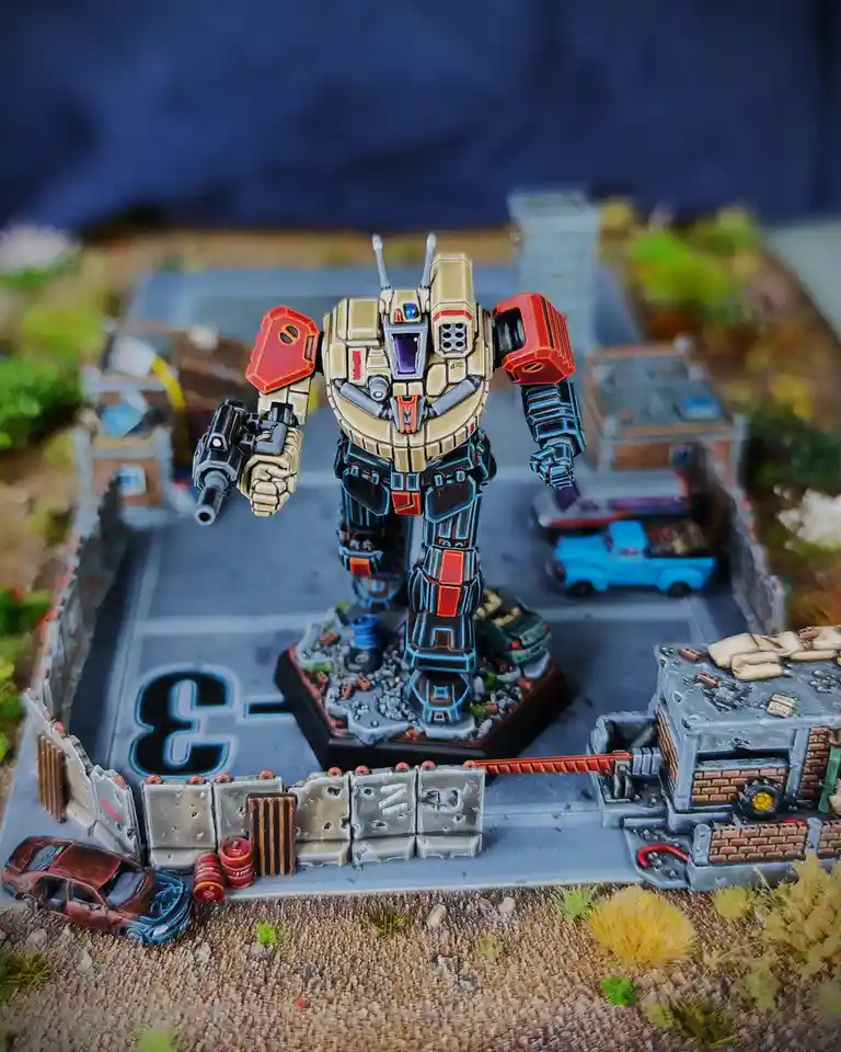 A painted miniature robot stands in a diorama of a ruined street with wrecked cars and buildings. The robot holds a weapon…