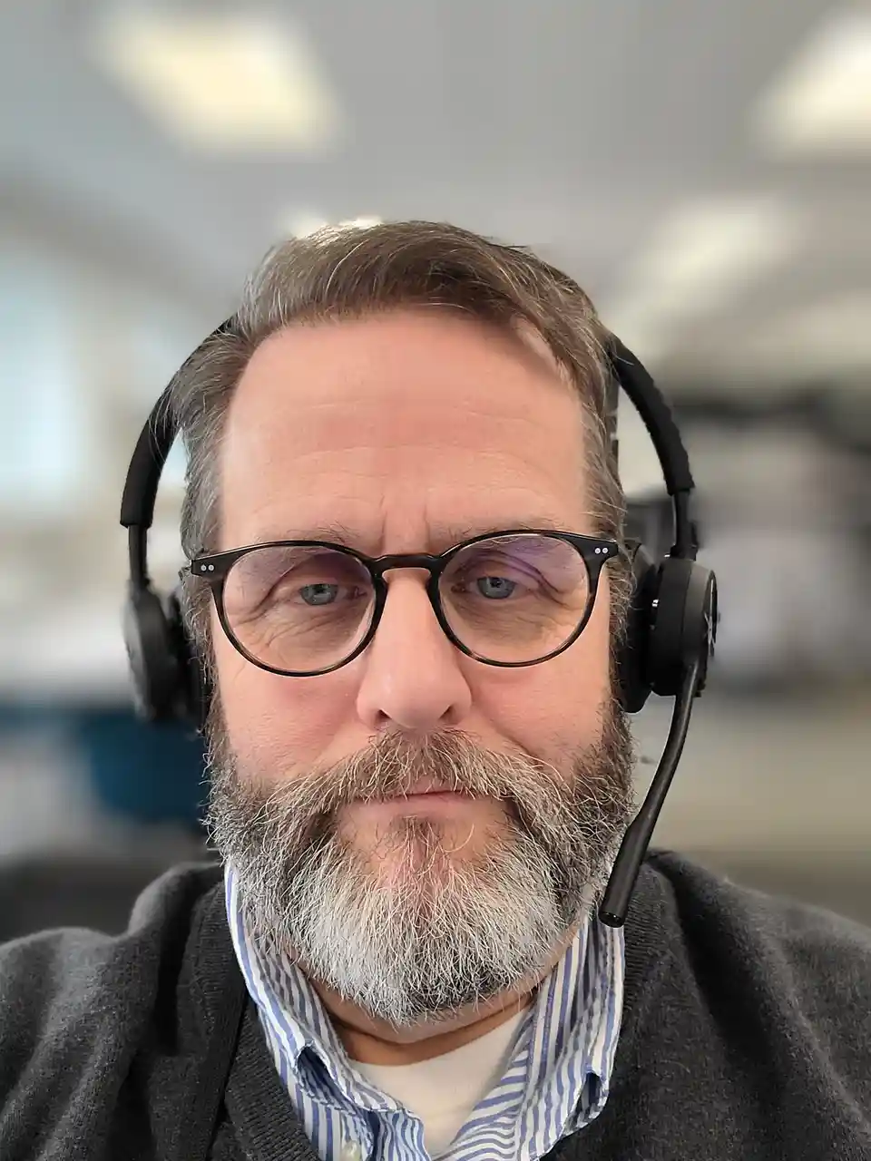 A close-up selfie of a middle-aged man with glasses and a gray beard, wearing a black headset with a microphone. He has a…