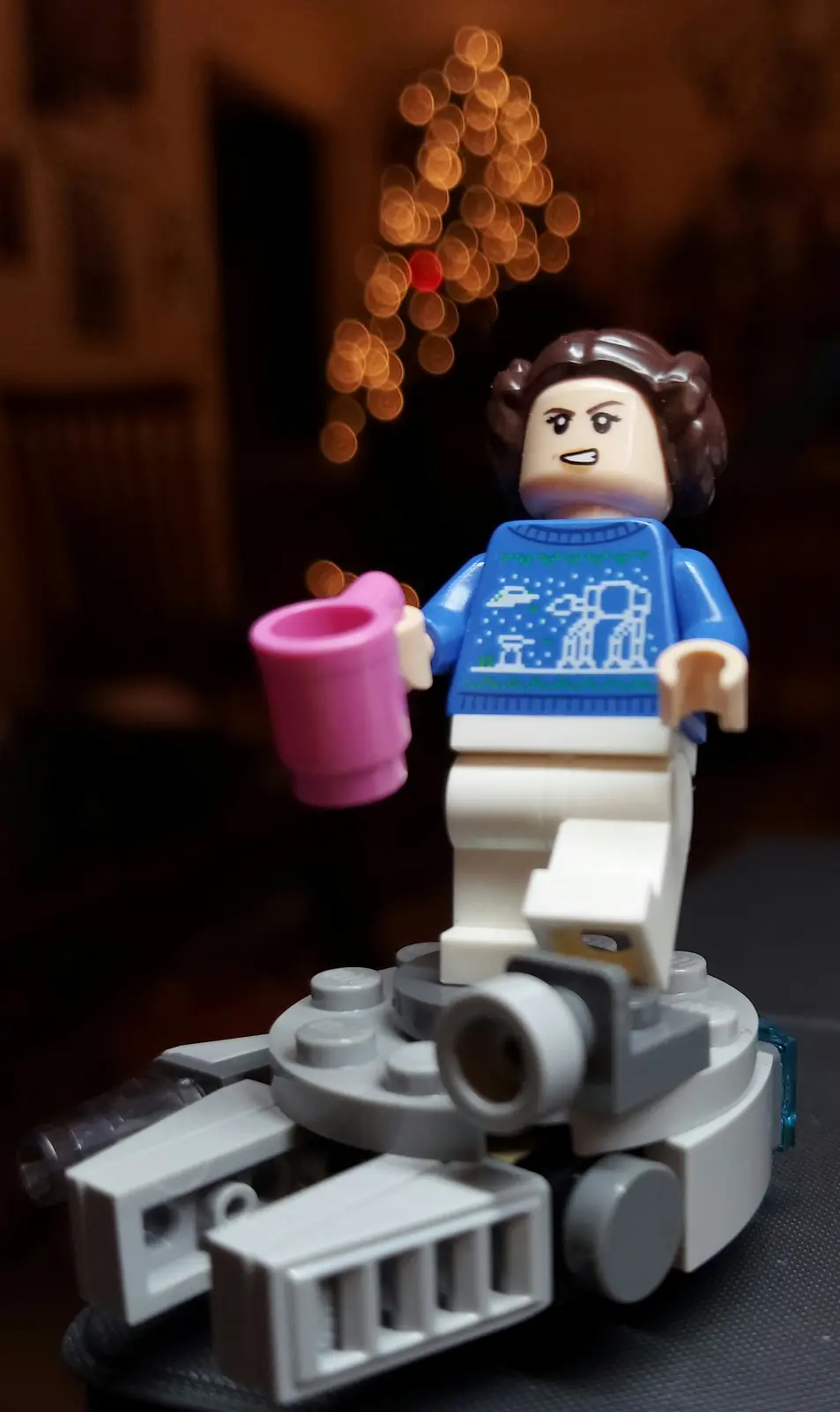 A lego minifigure resembling princess leia wears a blue christmas sweater with star wars characters and holds a pink mug…