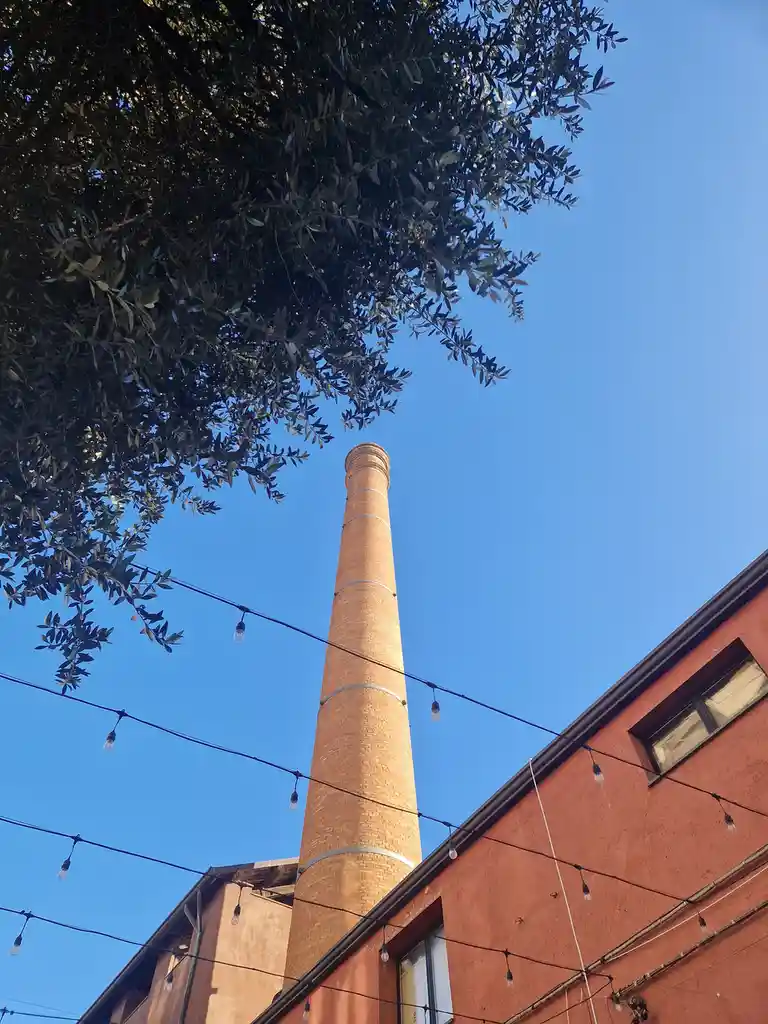 A low-angle photo shows a tall, brick industrial smokestack rising against a clear blue sky, framed by the dark green leaves…
