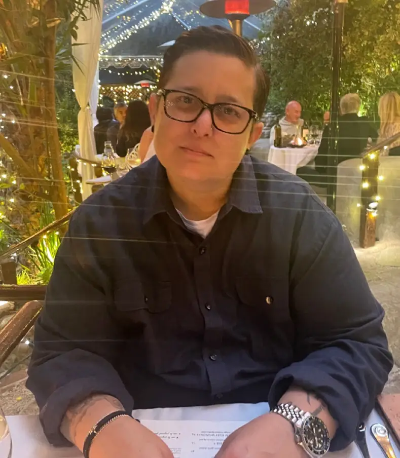 A person with glasses and a dark button-up shirt sits at an outdoor restaurant table, looking directly at the camera with a…