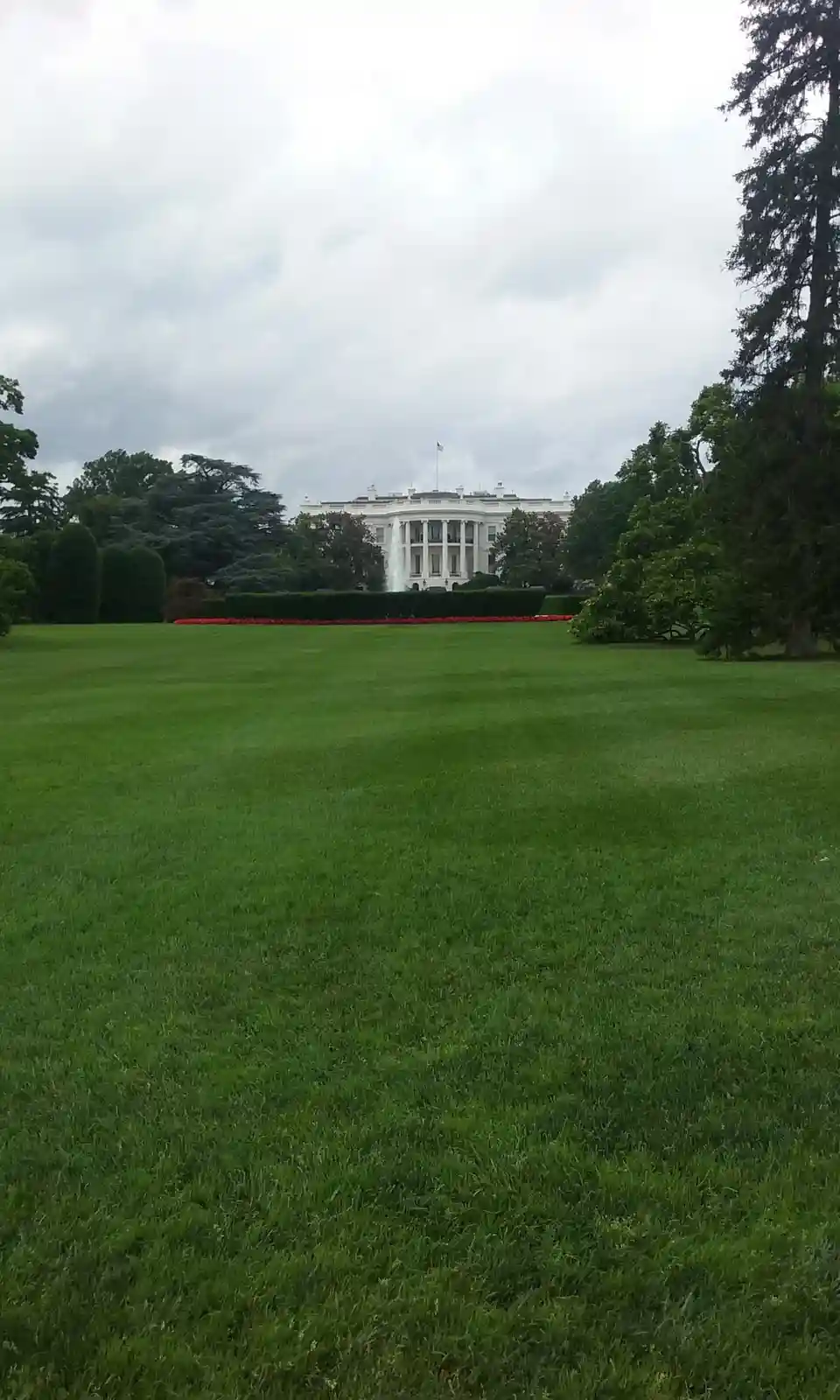 A wide-angle photo shows the White House in the distance, framed by a vast green lawn, trees, and a hedge with red flowers…