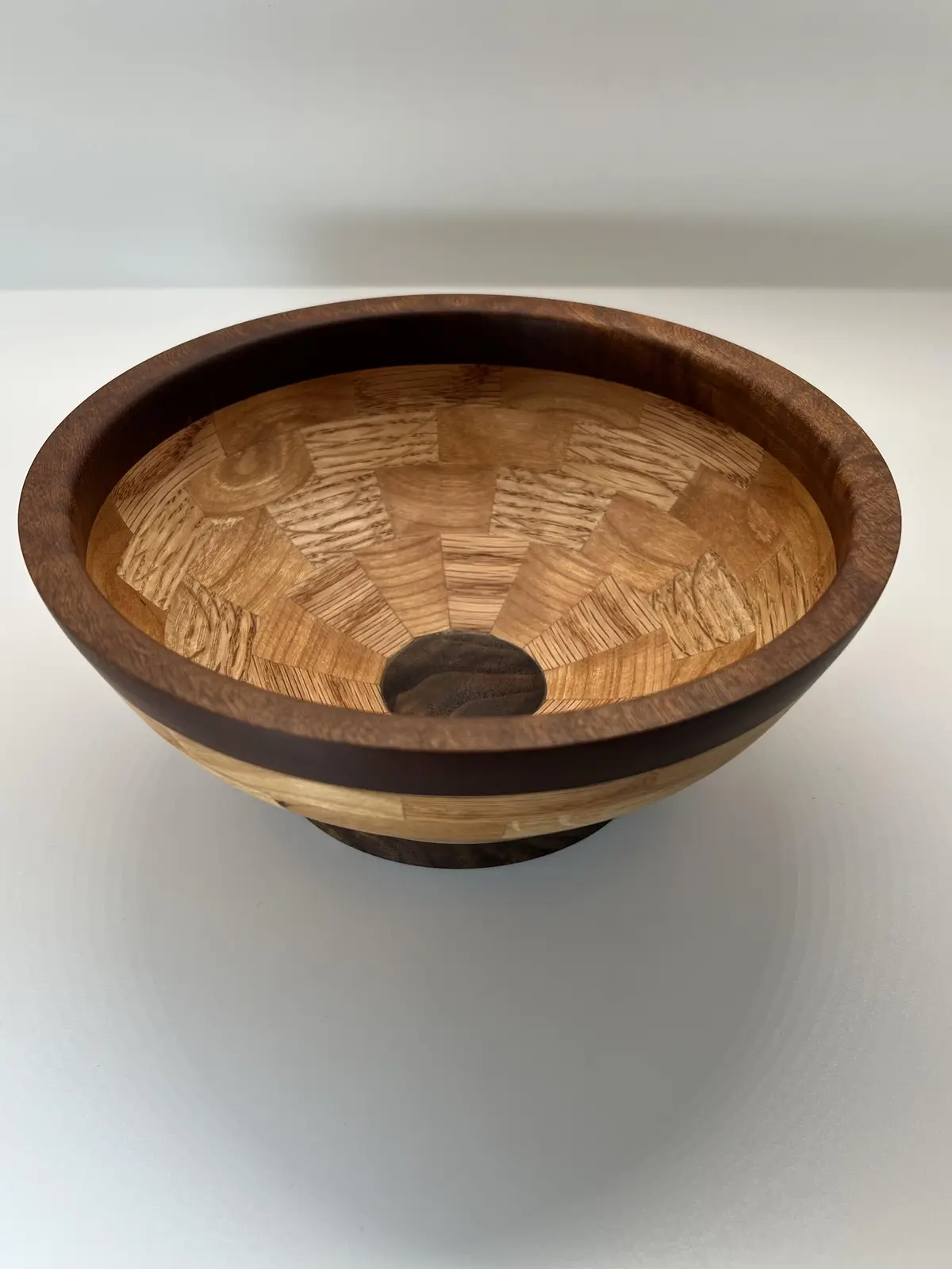 a highly polished wooden bowl with a geometric inlay pattern radiating from a dark circular center, resting on a white…